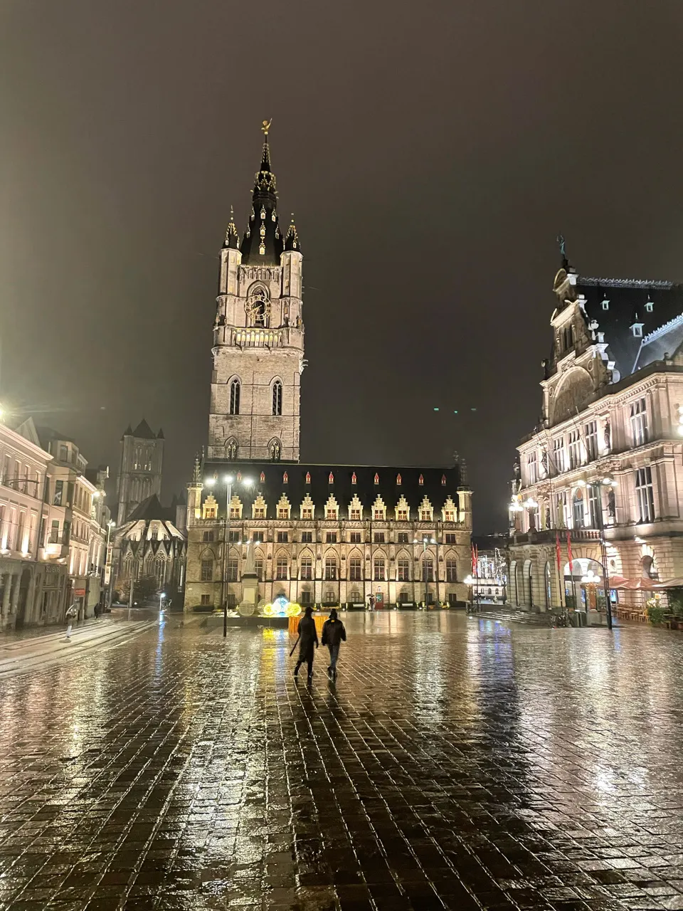 Het Belfort en het stadhuis van Gent aan een plein met natte kasseien in de avond