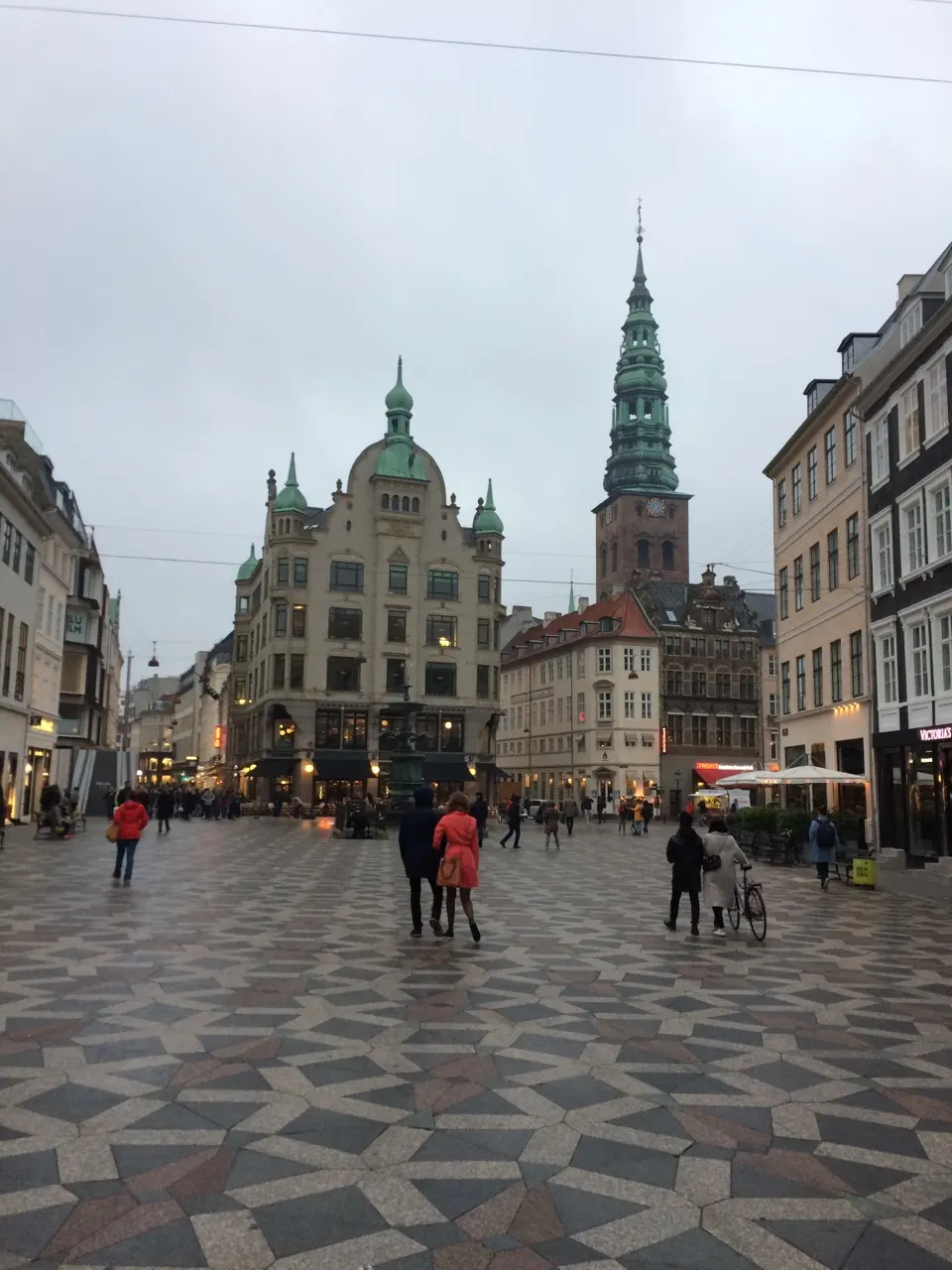 Een plein met geblokte bestrating en een kerktoren aan de winkelstraat Strøget